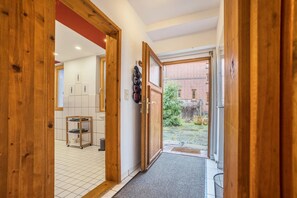 Un couloir avec une porte en bois donnant sur une pièce carrelée, une petite étagère avec des serviettes et une vue sur un bâtiment en briques à l’extérieur.