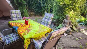 Ein Sitzbereich im Freien mit Tisch, bedeckt mit einer bunten Tischdecke, umgeben von Metallstühlen.