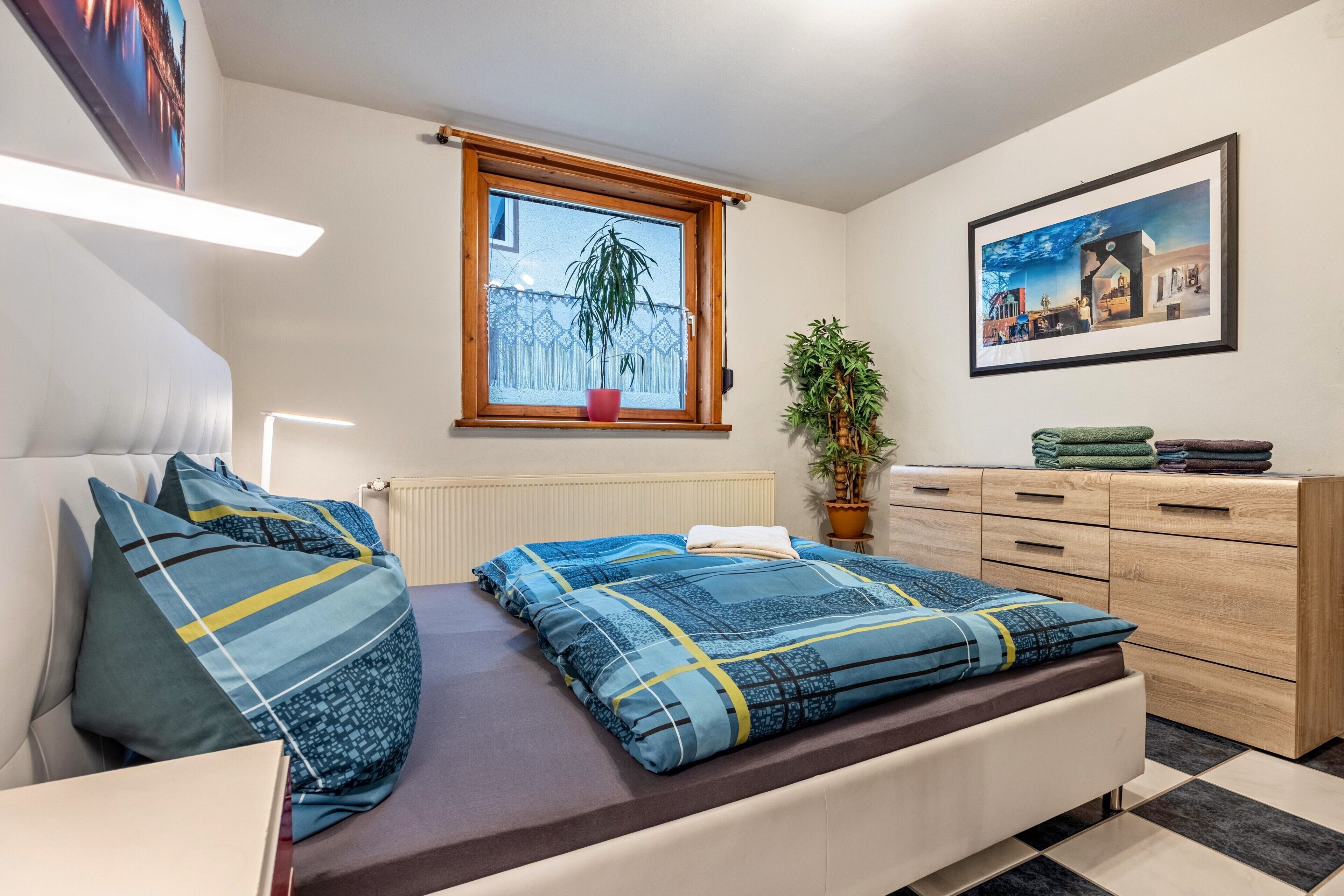 Ein Schlafzimmer mit Bett, Fenster, Heizkörper, Kommode und einem gerahmten Bild an der Wand.