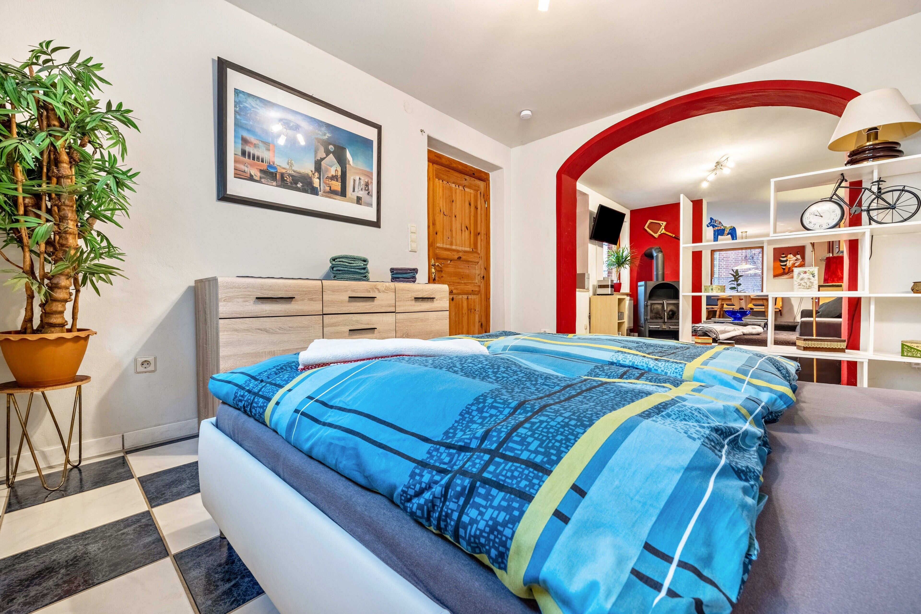 Een slaapkamer met een bed, een kamerplant, een houten dressoir, een rode boogvormige doorgang en een televisie.