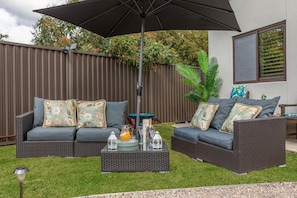 An outdoor seating area with two dark wicker sofas, each with floral cushions, a large umbrella, and a small table with drinks and a lantern.