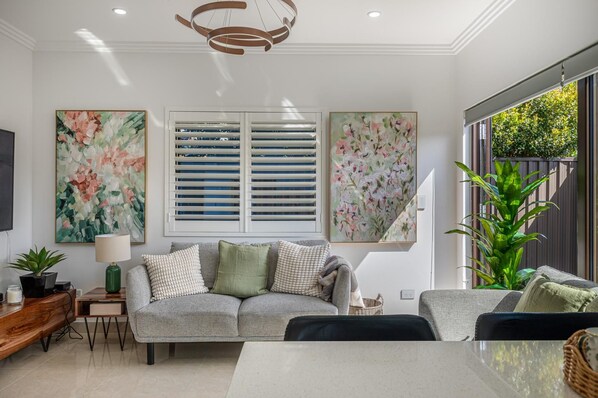 A modern living room with a grey sofa, a coffee table, and a TV on the left. There are two large floral paintings on the wall, a window with blinds, and a potted plant.