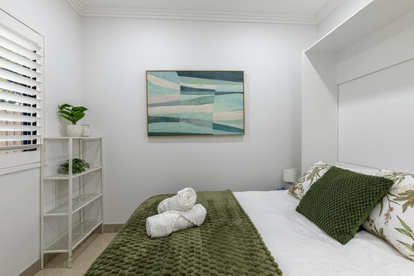 A bedroom with a bed, a green quilt, a white pillow, a green textured pillow, a wall-mounted abstract painting, a white shelf with a plant, and a white bedside table.