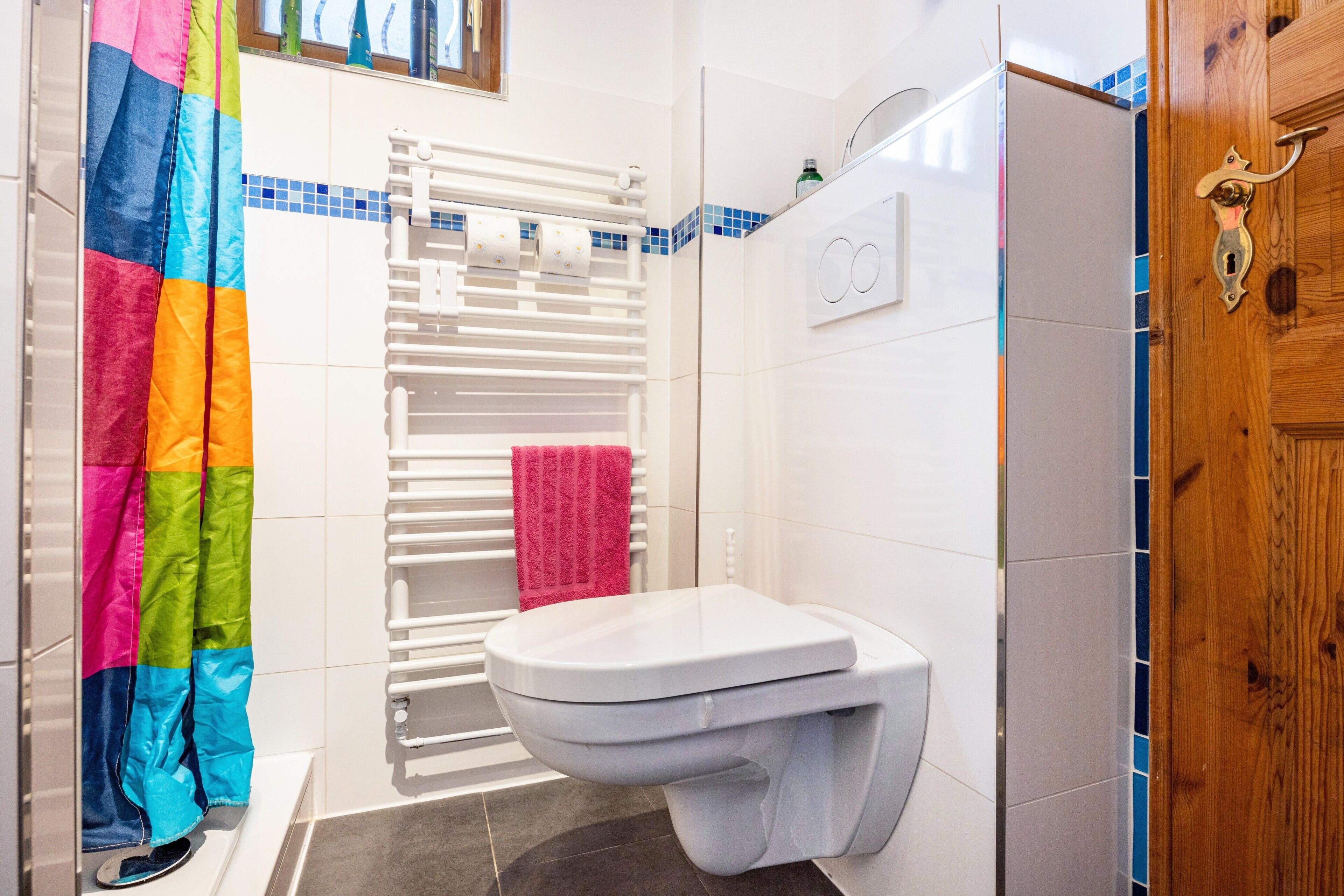 Un baño con inodoro blanco, ducha con puerta de madera y cortina colorida.