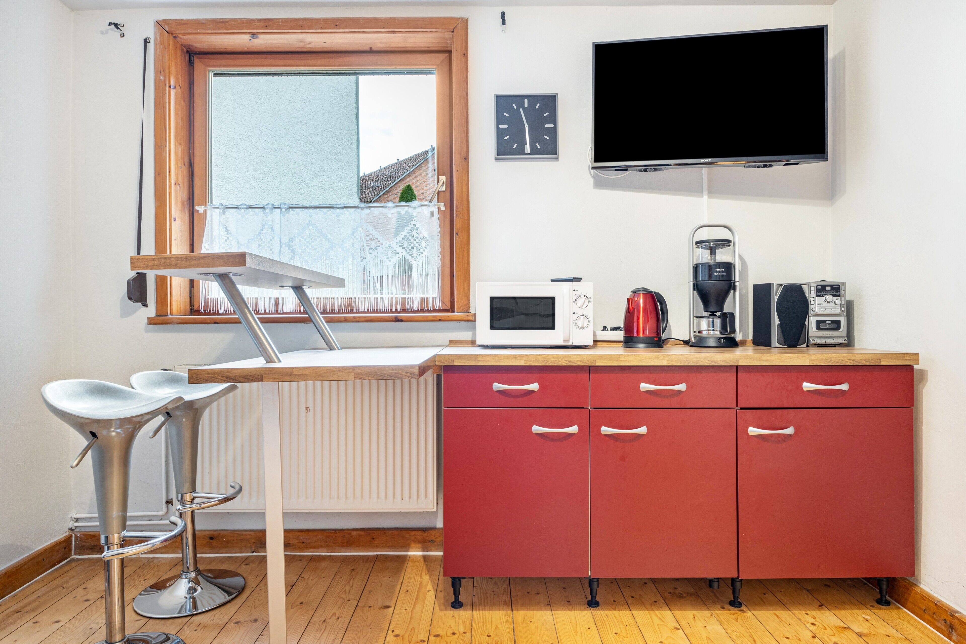 Eine Küche mit Holzarbeitsplatte, einem roten Schrank, einem Barhocker, einer Mikrowelle, einer Kaffeemaschine und einem an der Wand befestigten Fernseher.