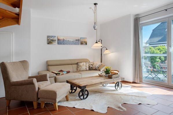 A living room with a sofa, armchair, wooden coffee table with wheels, a hanging lamp, and artwork on the wall.