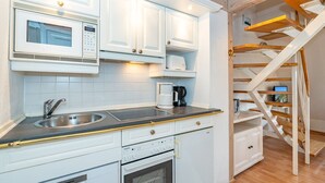 Una cocina moderna con armarios blancos, un lavamanos de acero inoxidable, un microondas Siemens y una cafetera. Se ve una escalera de madera con vista al área de estar.