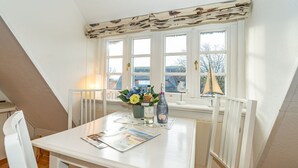 Una mesa de comedor blanca con un florero, dos botellas y revistas en una habitación con paredes blancas y techo inclinado.
