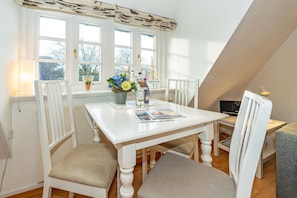 A dining area with a white table and chairs, a window with curtains, and a view of trees outside. - Altstadttor 10 - NEU AB 2024 in der Vermittlung !!! - Ferienwohnung für max 2. Personen nur 300m von (Sylt)