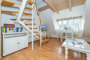 A spacious room with a wooden staircase, a dining area with white chairs, and a bookshelf filled with books. - Altstadttor 10 - NEU AB 2024 in der Vermittlung !!! - Ferienwohnung für max 2. Personen nur 300m von (Sylt)