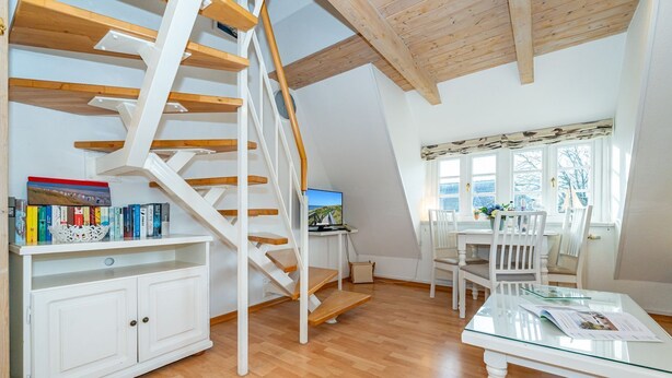 Una habitación amplia con una escalera de madera, un comedor con sillas blancas y una estantería llena de libros.
