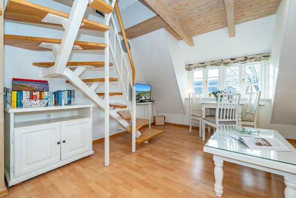 A spacious room with a wooden staircase, a dining area with white chairs, and a bookshelf filled with books.