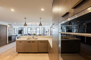 A modern kitchen with wooden cabinets, a large island, pendant lighting, and a visible clock.