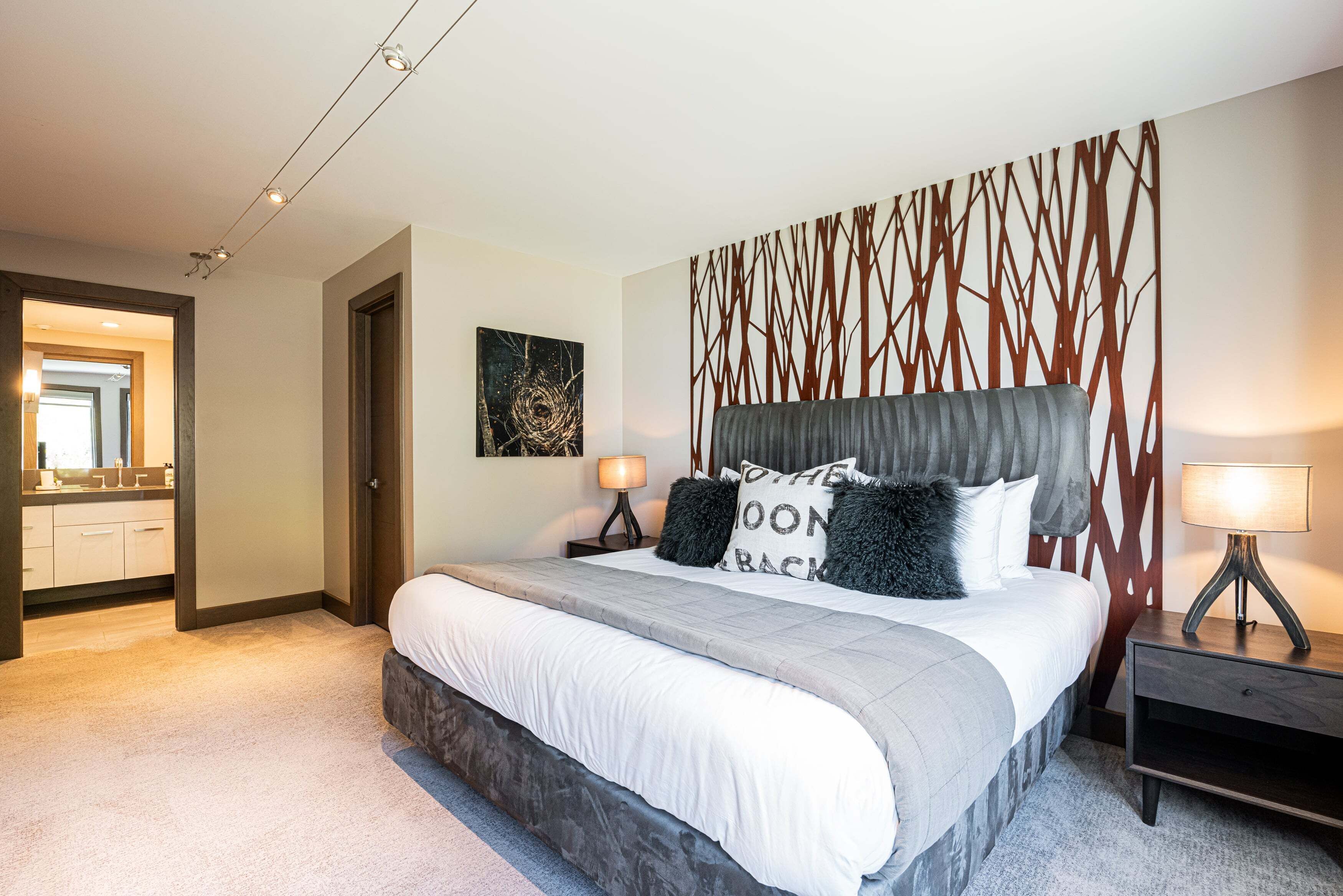 Une chambre à coucher comprenant un lit, des tables de chevet, une tête de lit ornée d’un motif géométrique, une œuvre d’art accrochée au mur, et une salle de bain visible à travers une porte ouverte.
