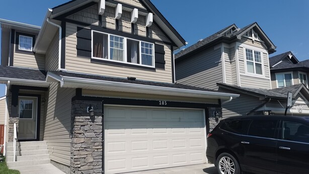 Une maison à deux étages avec une façade en pierre, un garage portant le numéro 285 et une voiture noire garée devant.