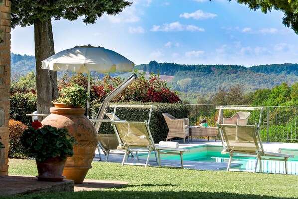 Uma área de piscina com espreguiçadeiras e um guarda-sol, cercada por vegetação e colinas ao fundo.