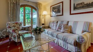 Una sala de estar acogedora con un sillón estampado floral, un sofá a rayas, una mesa de centro con diseño y un ventanal con cortinas.
