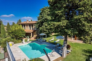 Uma área de piscina com espreguiçadeiras e guarda-sóis, cercada por um edifício de pedra e vegetação exuberante.