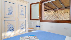 A room with a bed, a chair, and a stone wall. There are framed pictures on the wall and a window with a wooden frame.