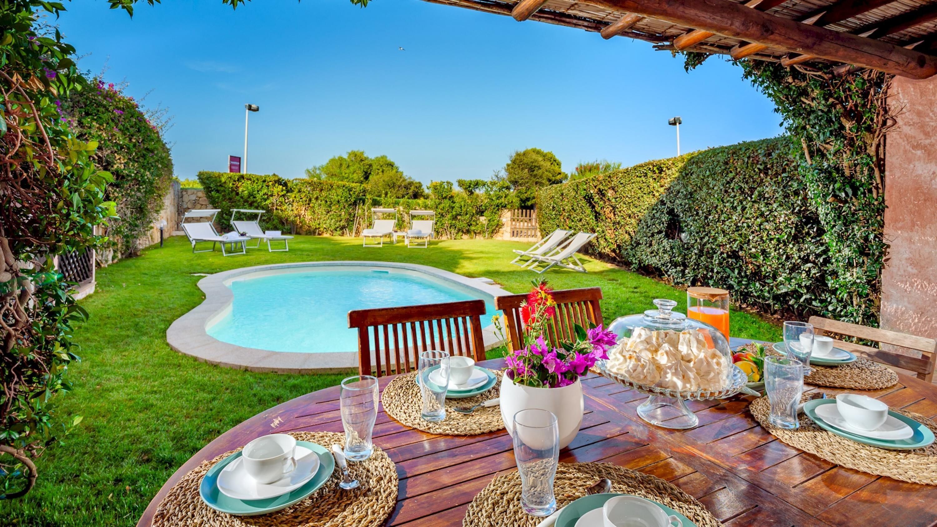 Uma área de refeições à beira da piscina, com uma mesa posta para dois, rodeada por vegetação exuberante e um céu azul límpido.