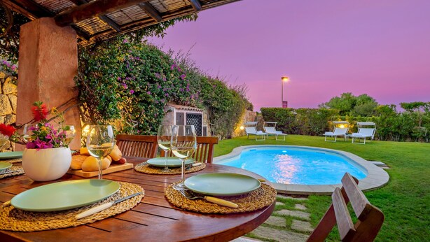 An outdoor dining area with a pool, surrounded by lush greenery and a stone wall.