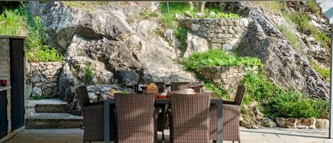 An outdoor patio with wicker furniture, a stone wall, and a rocky backdrop.