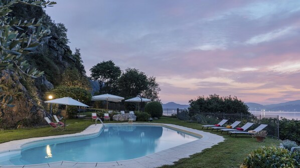 Piscina com espreguiçadeiras e guarda-sóis, cercada por vegetação e vista para um corpo d'água.