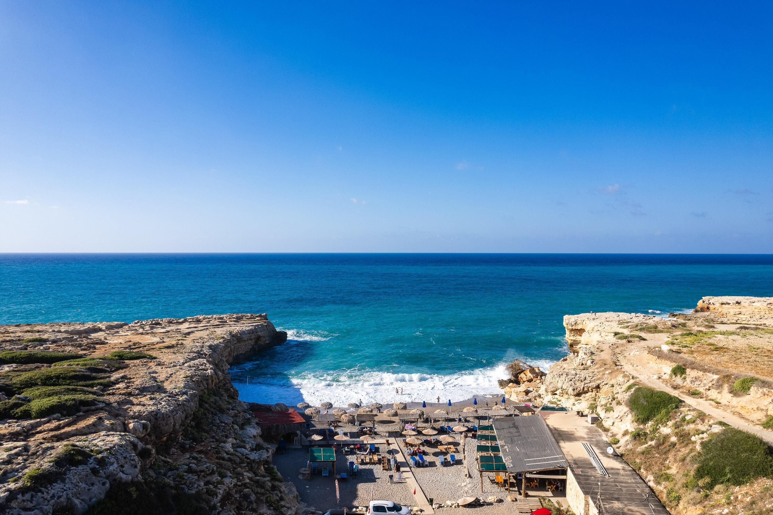 A coastal view with a rocky beach, a small beachside area with umbrellas, and a clear blue sky.