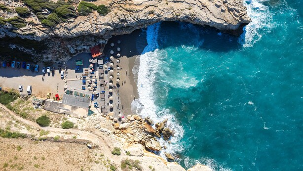 An aerial view of a coastal area with a beach, rocky cliffs, and a clear blue sea.