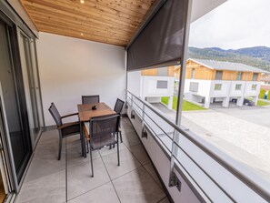 Un balcon avec une table et des chaises, une vue sur un immeuble aux façades en bois et un paysage montagneux en arrière-plan.
