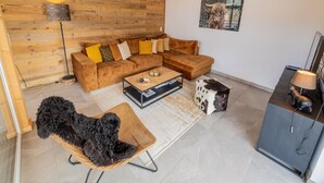 Una sala de estar moderna con una pared de acento de madera, un sofá modular con cojines estampados, una mesa de centro, un sillón negro de pelo y un reposapiés con estampado de vaca, y un televisor de pantalla plana sobre un soporte.
