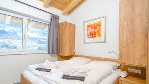 Un dormitorio con cama de madera, ventana con vista a un paisaje nevado, cortina gris y un cuadro en la pared.