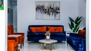 A lounge with a blue sofa, orange armchairs, a round table with a vase of flowers, and a large abstract painting on the wall. - Stratton 38 Asokoro (Abuja)