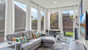 A modern living room with a grey sectional sofa, a coffee table, and a TV mounted on the wall. There is a dining area with a table and chairs visible through the large windows. - Red Mountain Villa, Game Room, Community Pool, Smart Tv's (Washington)