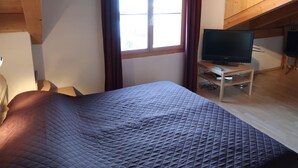 Une chambre à coucher avec un lit, un téléviseur posé sur un meuble, une fenêtre avec des rideaux et des poutres en bois au plafond.