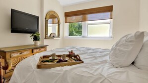 Une chambre d’hôtel avec un lit, une télévision, une commode en bois, une fenêtre donnant sur un paysage verdoyant et un plateau avec des tasses et une plante.