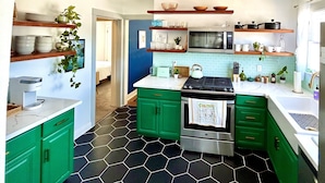 A modern kitchen with green cabinets, a stainless steel stove, and a microwave. There are shelves with dishes and plants, and a hexagonal tile floor.