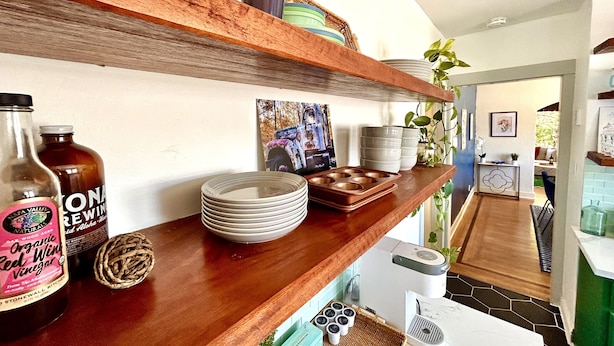 A kitchen with wooden shelves holding various items like a bottle of organic red wine vinegar, a coffee maker, and stacked plates.