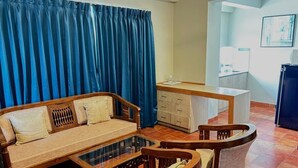 Sala de estar com sofá, mesa de centro e cadeiras, uma pequena cozinha e uma cortina azul.