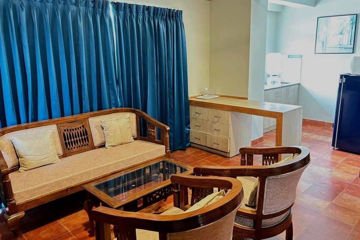 A living room with a sofa, coffee table, and chairs, a small kitchen area, and a blue curtain.