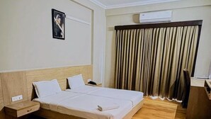 Quarto de hotel com piso de madeira, cama com roupa branca, mesa de cabeceira, escrivaninha, cadeira, janela com cortinas e ar condicionado.