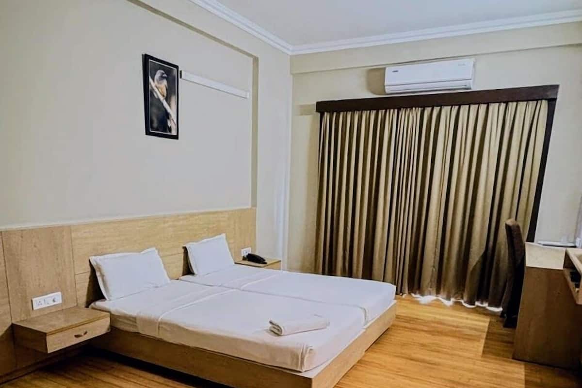 A hotel room with a wooden floor, a bed with white linens, a nightstand, a desk, a chair, a window with curtains, and an air conditioning unit.