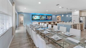 A modern kitchen with a central island, a dining area with glass tables, and a large TV mounted on the wall. - Arcade, Futuristic Theater, Triple 86” TV Wall, EV Charger, Waterpark - Epic Oasis (Davenport)