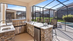 An outdoor kitchen with a grill, sink, and dishwasher, surrounded by a glass enclosure with a view of a backyard and a swimming pool. - Arcade, Futuristic Theater, Triple 86” TV Wall, EV Charger, Waterpark - Epic Oasis (Davenport)