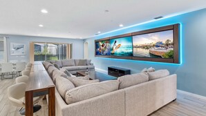 A modern living room with a sectional sofa, a long wooden table, and a large flat-screen TV mounted on the wall. - Arcade, Futuristic Theater, Triple 86” TV Wall, EV Charger, Waterpark - Epic Oasis (Davenport)