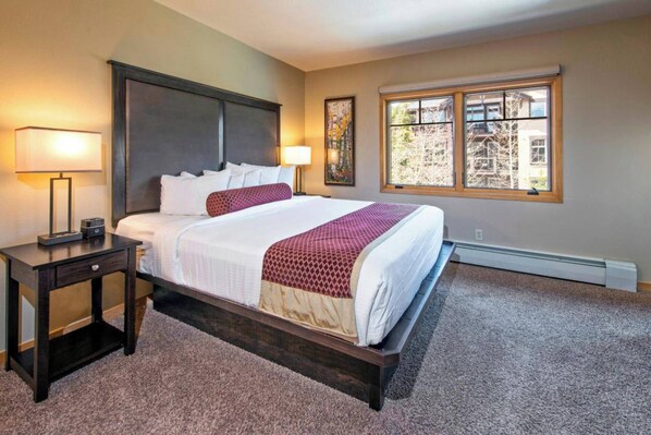 A bedroom with a large bed, two bedside lamps, a nightstand, a window with a view of trees, and a radiator.