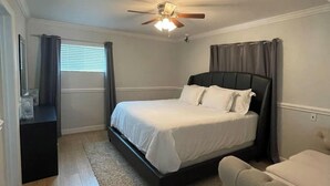 A bedroom with a bed, a ceiling fan, a window with curtains, a small table, and a chair.