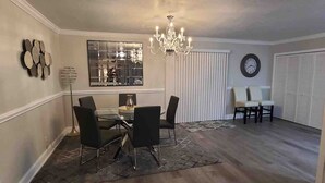 A dining area with a chandelier, a round table, and black chairs. There is a clock on the wall and a window with blinds.