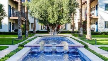 A swimming pool with a fountain, surrounded by palm trees and buildings.