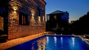 A swimming pool with stone tiles and a brick building with illuminated windows in the background. - Beautiful home in Cabrunici (Cabrunici)